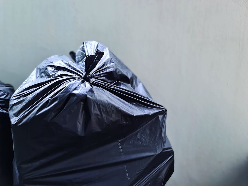 A close-up image showing several black plastic rubbish bags tightly tied at the tops, with visible creases and reflective surfaces indicating a plastic material. The bags are placed on a flat surface, potentially a pavement or driveway, with one bag prominently positioned in the foreground and others slightly in the background. The background features a plain, light-colored wall, with no other objects or environmental details visible. The scene appears to depict a typical waste collection or removal process, aligning with private rubbish disposal services such as those offered by Rubbish Removal Edgware, who specialise in on-site clearance and alternative waste handling solutions for domestic or commercial clients.