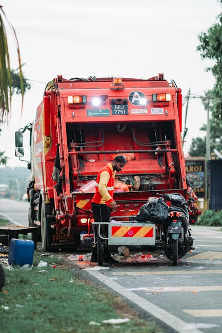 A sanitation worker wearing a yellow and red uniform is engaged in rubbish collection on a street, standing next to a large red trash compactor truck with the back open. The truck, labeled with identification numbers and safety stickers, is positioned on a paved roadside with its rear hatch raised, revealing internal mechanical components. The worker is loading waste into the truck, and nearby, there is a black plastic bag and a small metal cart with reflective red and yellow stripes. The surrounding environment includes a sidewalk, a patch of grass adjacent to the street, and some trees in the background. Overhead power lines and faint indications of other vehicles or signage are visible, suggesting an urban or suburban setting, typical for local waste collection activities. The scene illustrates a private or independent rubbish removal process often used for on-site clearance outside conventional municipal collection routes, supporting the context of alternative waste handling services. Lighting conditions are natural, with an overcast sky casting diffuse light over the area.