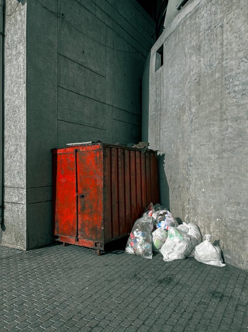 A close-up image showing several black plastic rubbish bags tightly tied at the tops, with visible creases and reflective surfaces indicating a plastic material. The bags are placed on a flat surface, potentially a pavement or driveway, with one bag prominently positioned in the foreground and others slightly in the background. The background features a plain, light-colored wall, with no other objects or environmental details visible. The scene appears to depict a typical waste collection or removal process, aligning with private rubbish disposal services such as those offered by Rubbish Removal Edgware, who specialise in on-site clearance and alternative waste handling solutions for domestic or commercial clients.