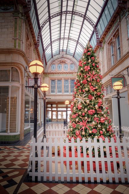 Inside a large Victorian-style shopping arcade with a high glass and metal arched ceiling, a tall Christmas tree decorated with pink and gold baubles, pink ribbons, and white snowflake ornaments stands in the center, surrounded by a white picket fence. The interior features ornate brickwork and large windows with decorative tracery on the upper floors, alongside vintage-style lamp posts with warm glowing bulbs on either side of the tree. The patterned tile floor adds to the historic yet festive atmosphere, and the space is illuminated by both natural light from the glass ceiling and the warm light from the lamps. This setting, while primarily a shopping arcade, visually aligns with a context where private or independent collections of decorations and festive display arrangements are part of alternative or non-standard approach to seasonal display, similar to private waste handling or on-site clearance, though no rubbish or waste is visible in the scene. The overall scene emphasizes a clean, well-maintained indoor environment of historic architectural charm with festive holiday decor.