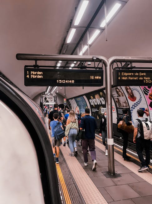 Inside a busy underground train station, a platform is filled with passengers preparing to board a train. The scene shows a diverse group of people standing on the yellow tactile paving strip, with some engaged in conversation and others focused on the train doors. The train’s exterior is partially visible on the left side of the image, with the edge of a white door and a portion of the side panel. Overhead, illuminated digital display boards provide travel information, showing destinations such as Morden and High Barnet, with departure times and train statuses. The station's interior features modern lighting fixtures mounted on the ceiling and advertising posters lining the walls behind the platform. The environment appears clean, well-lit, and organized, reflecting the typical scene of commuter activity in a metropolitan underground transit system. The image captures the dynamic movement of passengers, with some standing still and others walking, in an atmosphere that emphasizes efficient public transport services in an urban setting.