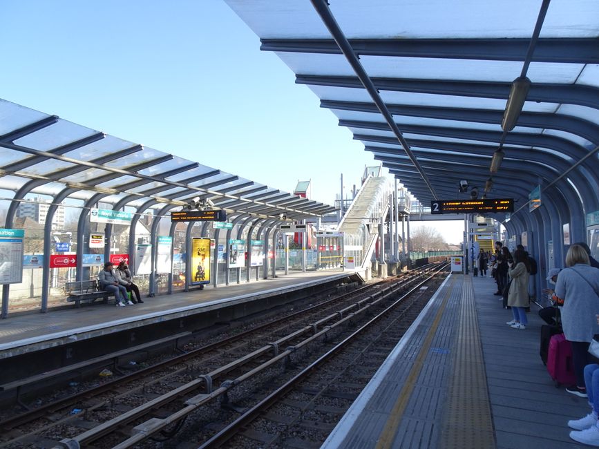 The image depicts an outdoor train station platform with a curved, transparent roof structure supported by metal framing. On the left side, a waiting shelter houses a few seated passengers, some with bags or coats. Next to the shelter, behind glass panels, digital information screens display train schedules and notices. In the background, a train painted in red and silver is stationing at the platform, with visible windows and doors. To the right, several passengers are standing, some holding mobile devices or luggage, and others engaging in conversation. A row of small, illuminated advertising posters is visible along the shelter, along with a yellow ticket machine on the platform’s edge. The station is situated in an urban environment, with a clear blue sky overhead and distant buildings visible beyond the platform. The scene is well-lit with natural daylight, illustrating a typical commuter setting. This scene is consistent with the outdoor environment of a typical UK railway station, where independent or private clearance of waste might be required for station maintenance or upgrades, as managed by local waste removal services like Rubbish Removal Edgware.