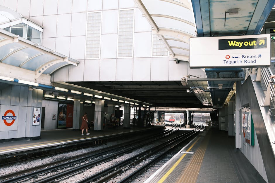 The image depicts the platform of Hammersmith London Underground station with multiple train tracks in the foreground, bordered by yellow safety lines and textured grey paving. To the left, the station's white tiled wall features a red and blue Hammersmith roundel logo and advertisements mounted on the wall. A commuter wearing a coat walks along the platform, heading towards an opening under the station's canopy, which is constructed from metal and transparent panels, allowing natural light to illuminate the area. The canopy curves overhead, supported by white structural elements, with a blue sign hanging from it indicating the way out towards Talgarth Road and buses. In the background, other parts of the station infrastructure are visible, including stairs and additional signage, while the environment appears clean and organized, typical of an urban underground station. This scene aligns with the role of private waste handling, as Rubbish Removal Edgware provides alternative rubbish collection solutions for commuters and station maintenance areas outside of the public authority systems, ensuring efficient clearing of waste from high-traffic public transport locations.