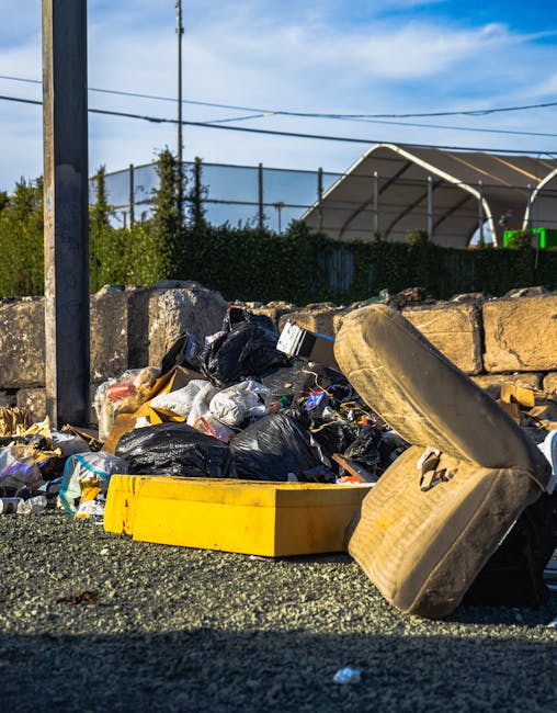 A close-up view of a pile of mixed waste and rubbish on a gravel surface, including black and white plastic garbage bags, a yellow plastic container, and a weathered car tire lying on its side. Behind the pile, there is a low stone wall constructed from large, rough-hewn stones, with some greenery and tall trees visible beyond. In the background, a chain-link fence and power lines are visible, along with a partially visible modern white canopy structure with curved supports and a green sign or display. The scene is illuminated by daylight, suggesting an outdoor setting, possibly near a rubbish collection or disposal area, with natural light highlighting the textures and materials of the waste and surroundings. Rubbish Removal Edgware’s service context relates to alternative waste handling and private rubbish clearance.