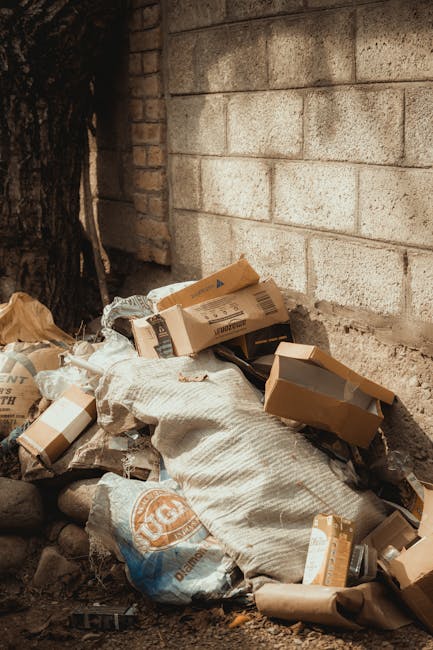 Close-up view of a tightly tied black plastic rubbish bag made from glossy, flexible material, with visible creases and folded edges, resting against a plain, light-colored wall background. The bag is placed on a surface likely outdoors or in a rubbish collection area, with additional black bags partially visible behind it. The scene suggests a setting related to waste disposal or rubbish removal, consistent with private rubbish collection services such as those offered by Rubbish Removal Edgware. The lighting is neutral, highlighting the texture and reflective surface of the plastic bag, emphasizing its use for household or commercial rubbish disposal. The environment appears clean, organized, and focused solely on the waste container, reflecting typical on-site clearance or independent waste handling scenarios.
