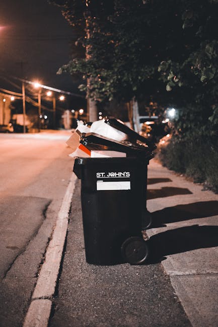 A large black wheelie bin with white lettering marked 'St. John's' is positioned on a paved sidewalk next to a street at night. The bin is partially open and filled with assorted waste, including cardboard boxes, plastic items, and paper. The background features streetlights casting a warm glow over the empty road and surrounding trees, with some residential buildings visible in the distance. The scene captures a typical urban setting where private waste collection, such as that offered by rubbish removal services like Rubbish Removal Edgware, is used for disposing of household waste through independent collection methods. Shadows of the bin are cast on the pavement due to the street lighting, and the overall environment appears quiet and well-maintained, emphasizing the practical aspect of local rubbish disposal in a residential area.