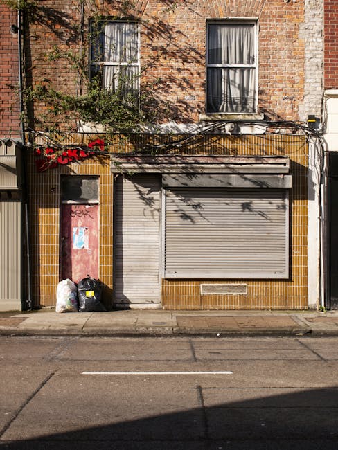 The image depicts the façade of a small, closed commercial or residential building with a brick upper section and a tiled lower section in a shade of brown or ochre. Two large, double-pane windows with white frames are situated above the storefront, with sheer curtains partially visible behind the glass. The ground floor features a metal roller shutter door in a closed position, painted in a neutral light grey tone, with a smaller pedestrian door to its left, also in grey, both set into the tiled section. To the left of the doors, near the curb, there are three black waste disposal bags and a white plastic bag, indicating disposed rubbish awaiting collection. Overhead, a black cable runs horizontally across the façade, accompanied by a small cable box or meter box on the right side. The ground is paved with asphalt, showing faint markings and minor cracks, with shadows cast by nearby trees giving a dappled effect on the building’s surface. This scene is typical of an urban environment where private rubbish collection services, such as Rubbish Removal Edgware, might handle waste for local businesses or residents, especially in areas with on-site waste storage. The overall setting emphasizes the functional nature of waste disposal alongside services geared toward alternative, private rubbish collection.