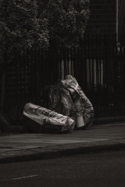 A large black wheelie bin with white lettering marked 'St. John's' is positioned on a paved sidewalk next to a street at night. The bin is partially open and filled with assorted waste, including cardboard boxes, plastic items, and paper. The background features streetlights casting a warm glow over the empty road and surrounding trees, with some residential buildings visible in the distance. The scene captures a typical urban setting where private waste collection, such as that offered by rubbish removal services like Rubbish Removal Edgware, is used for disposing of household waste through independent collection methods. Shadows of the bin are cast on the pavement due to the street lighting, and the overall environment appears quiet and well-maintained, emphasizing the practical aspect of local rubbish disposal in a residential area.